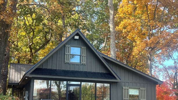 Une cabane moderne dotée d’une grande façade vitrée, d’un espace extérieur aménagé et entourée d’arbres aux couleurs automnales.