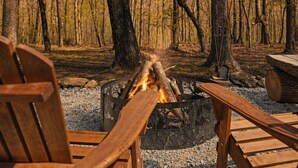 Ein Lagerfeuer mit Holzscheiten, umgeben von Holzstühlen und einer Bank, inmitten eines bewaldeten Gebiets.