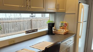 Eine Küche mit Esstisch, zwei Stühlen, einer Kaffeemaschine, einem Kühlschrank und einem Fenster mit Blick auf einen Holzzaun.
