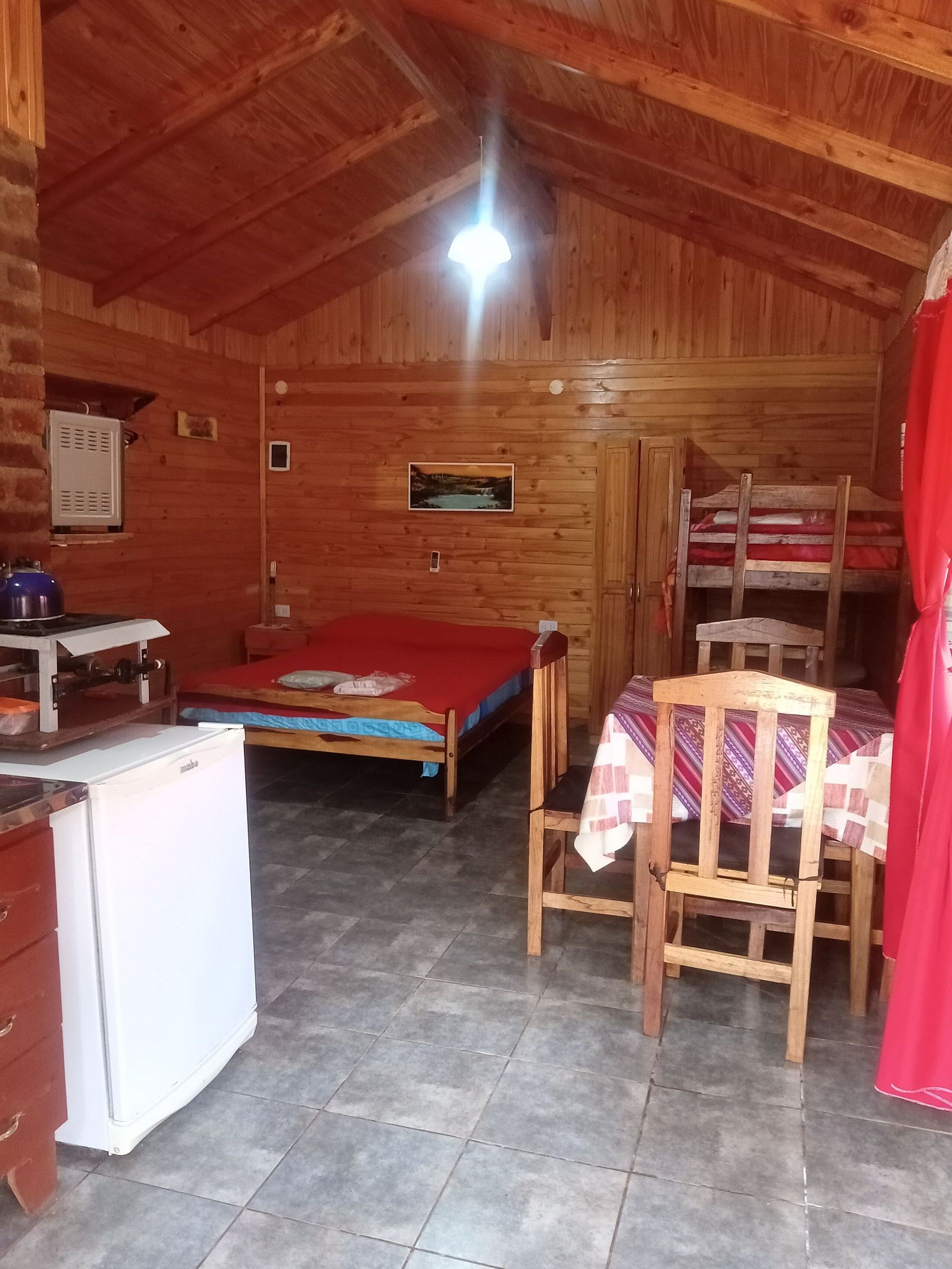 A wooden cabin interior with a bed, a table with a chair, a refrigerator, and a wooden bunk bed.