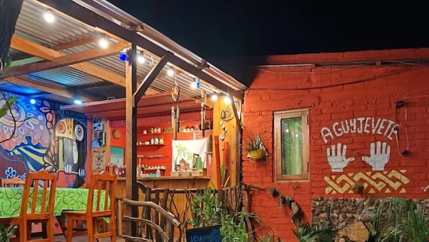 An outdoor seating area with wooden chairs and tables, a chalkboard sign, and a red building with a handprint mural. - Bungalows El Pindó (Colonia Alberdi)