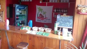 A bar area with a red wall, a sign for 'BUNGALOWS EL PINDO', a refrigerator with beverages, and a chalkboard menu. - Bungalows El Pindó (Colonia Alberdi)