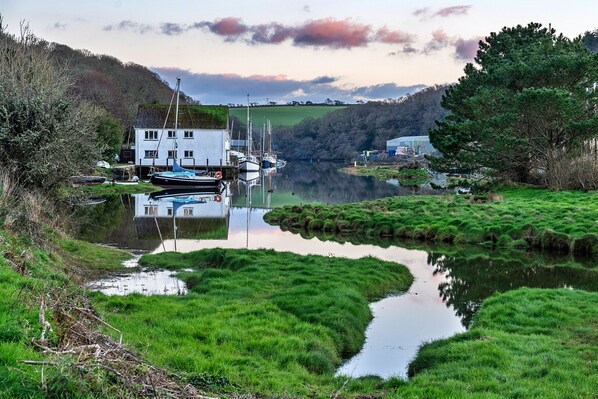 A riverside view with boats docked, a white building, and a forested hill in the background. - The Old Counting House (Gweek)