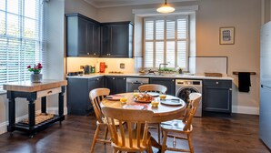 A kitchen with dark cabinets, a wooden dining table, and chairs. There is a window with blinds, a washing machine, and a sink.