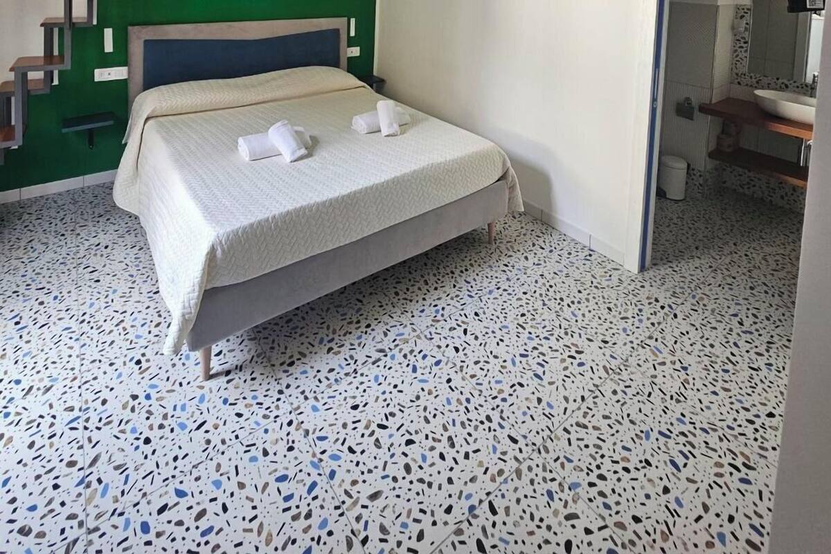 Un dormitorio con cama, paredes verdes, una escalera y un baño visible a través de una puerta abierta.