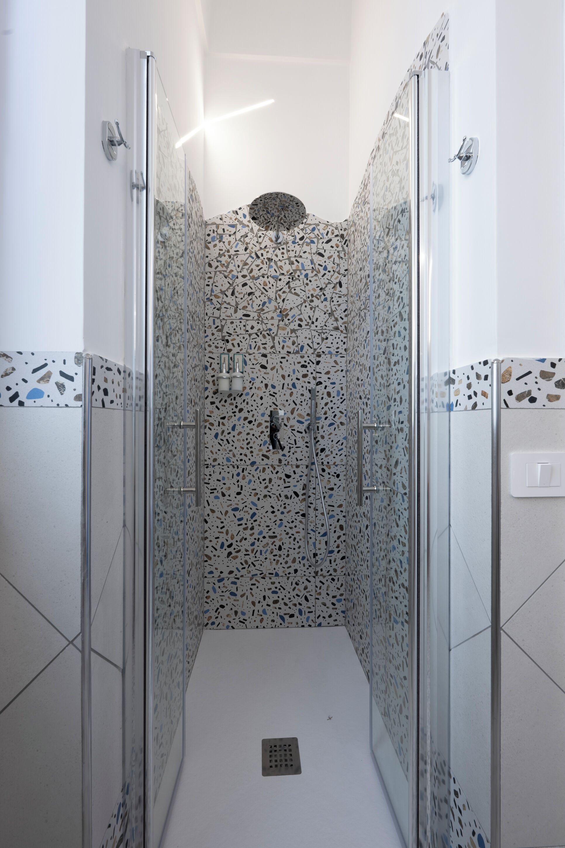Una ducha con cerramiento de vidrio, pared de azulejos con vetas y suelo blanco.