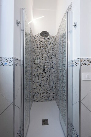 Una ducha con cerramiento de vidrio, pared de azulejos con vetas y suelo blanco.
