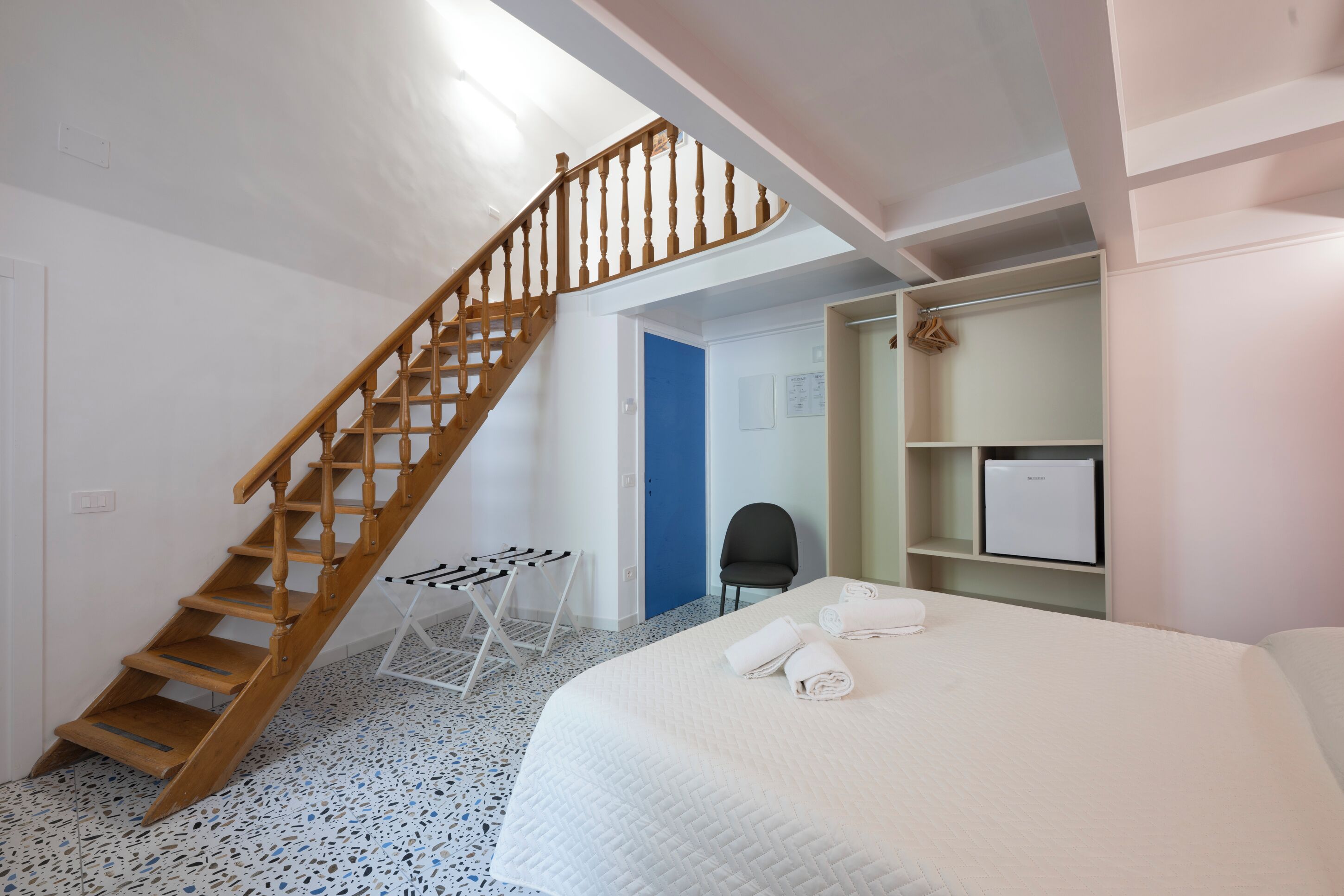 Un dormitorio con una escalera de madera, una cama con ropa blanca, una mesita pequeña y una silla.