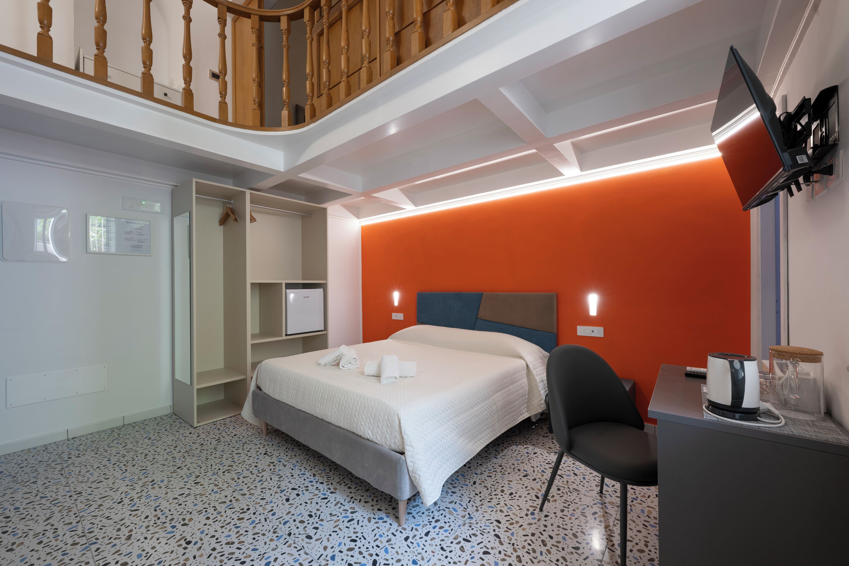 A hotel room with a bed, a desk, a chair, a TV mounted on the wall, and a staircase.