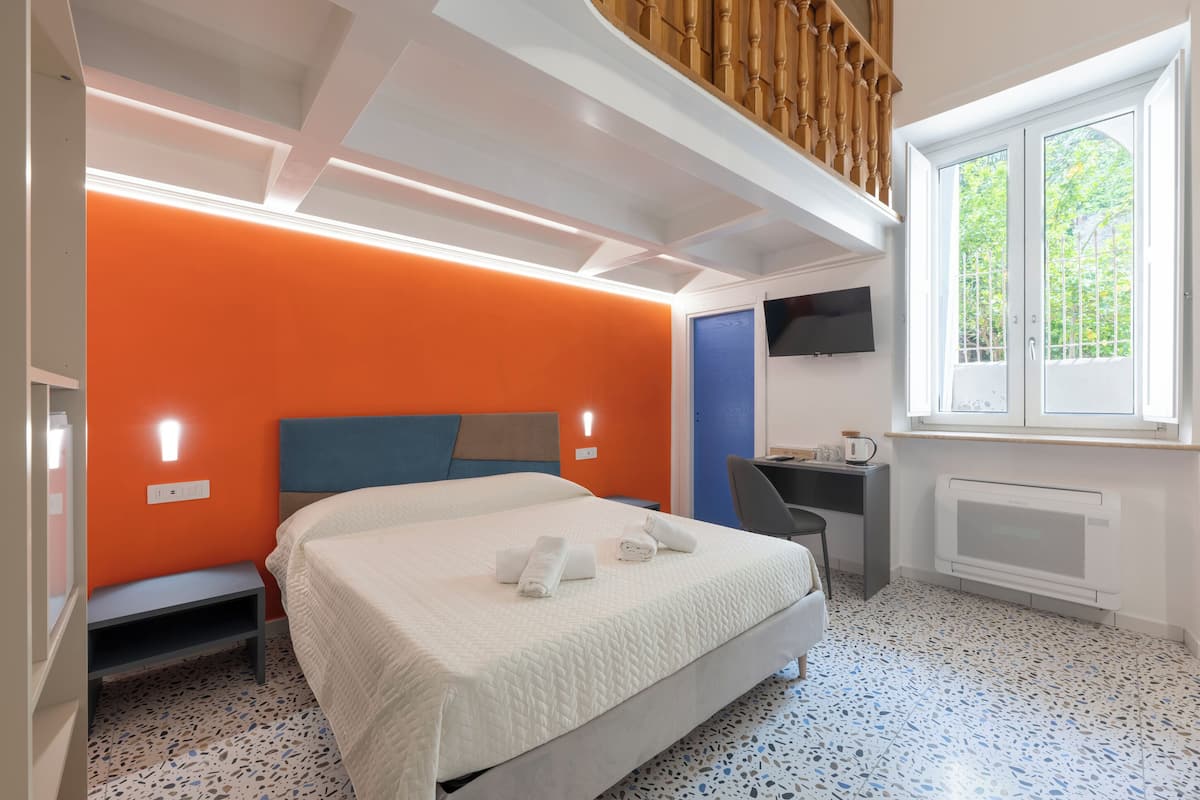 Un dormitorio con cama, escritorio, silla, televisor y ventana con vista a árboles.