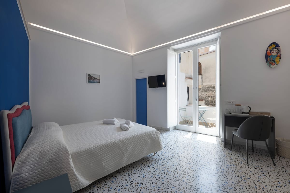 Un dormitorio con cama, escritorio y silla. Hay una puerta que da a un balcón.