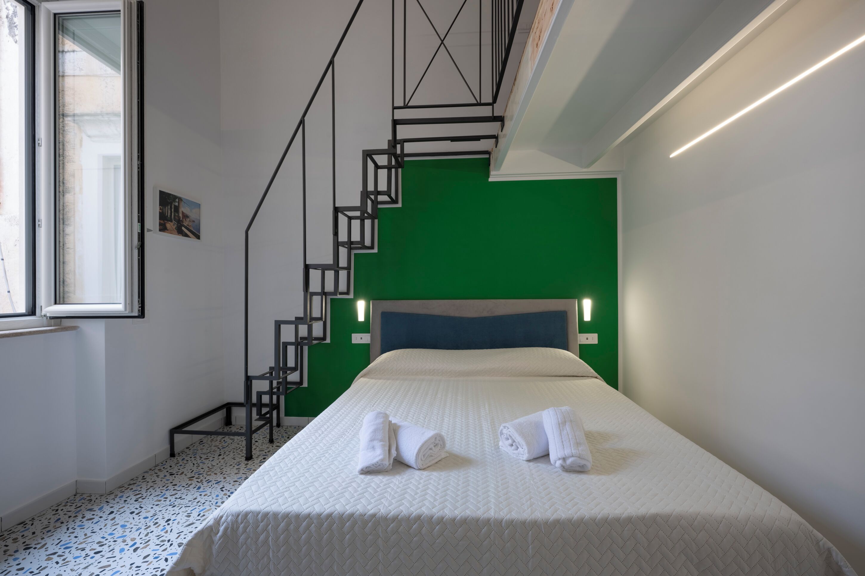 Una habitación de hotel con cama, pared de acento verde, escalera y ventana.