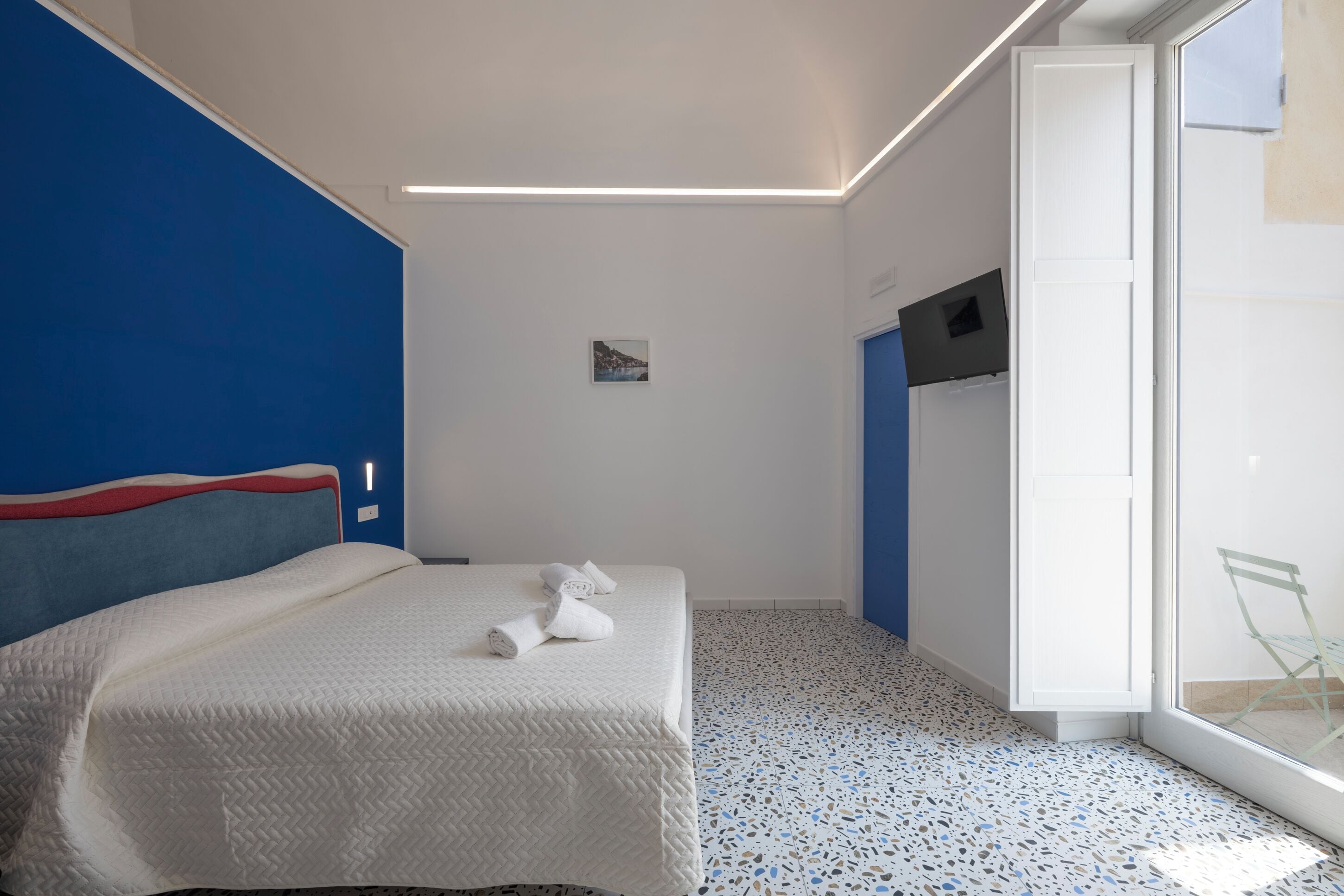 A modern hotel room with a bed, a small bedside table, a TV mounted on the wall, and a glass door leading to a balcony.