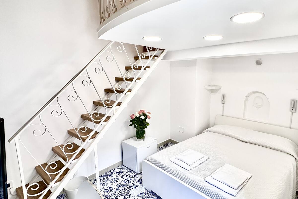 Un dormitorio con una cama blanca, una mesita de noche blanca y una escalera curva de madera con barandillas blancas, además de un suelo con estampado azul y blanco.