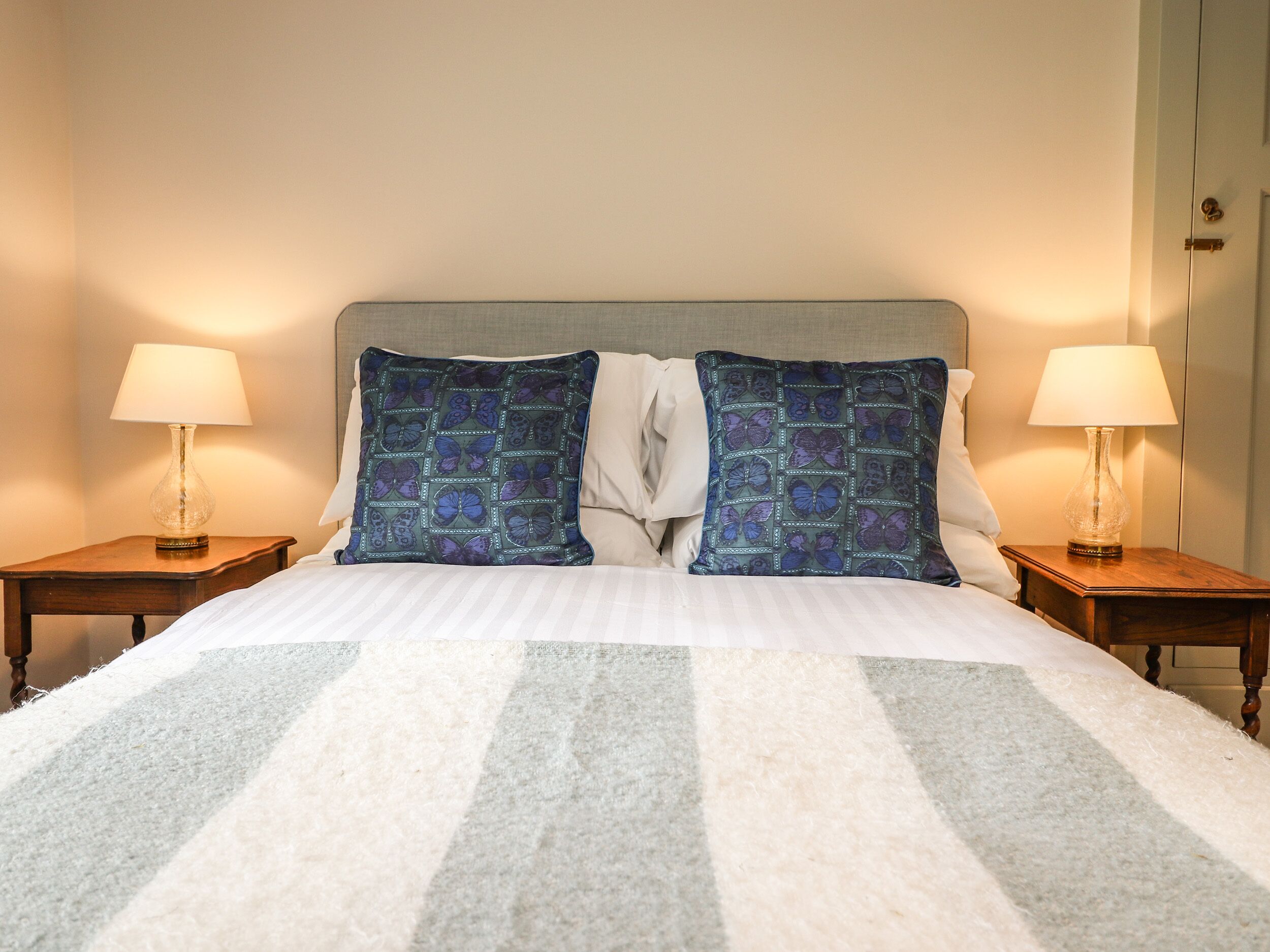 A bed with a striped blanket, two square pillows with a butterfly pattern, and two bedside lamps on wooden nightstands.