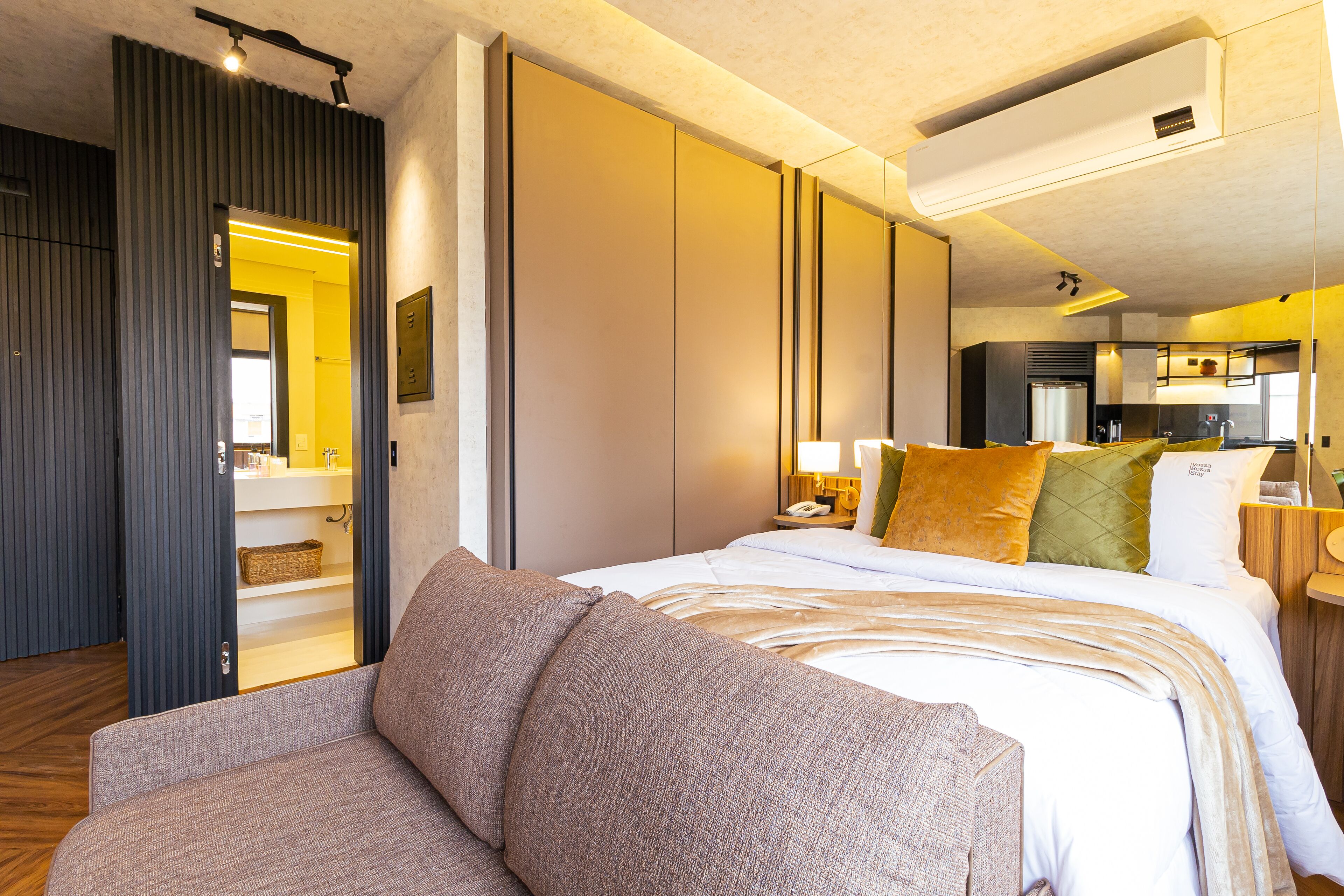 Habitación de hotel moderna con una cama grande, un sofá, una zona de cocina y un baño visible a través de una puerta abierta.