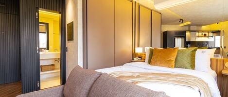 Habitación de hotel moderna con una cama grande, un sofá, una zona de cocina y un baño visible a través de una puerta abierta.