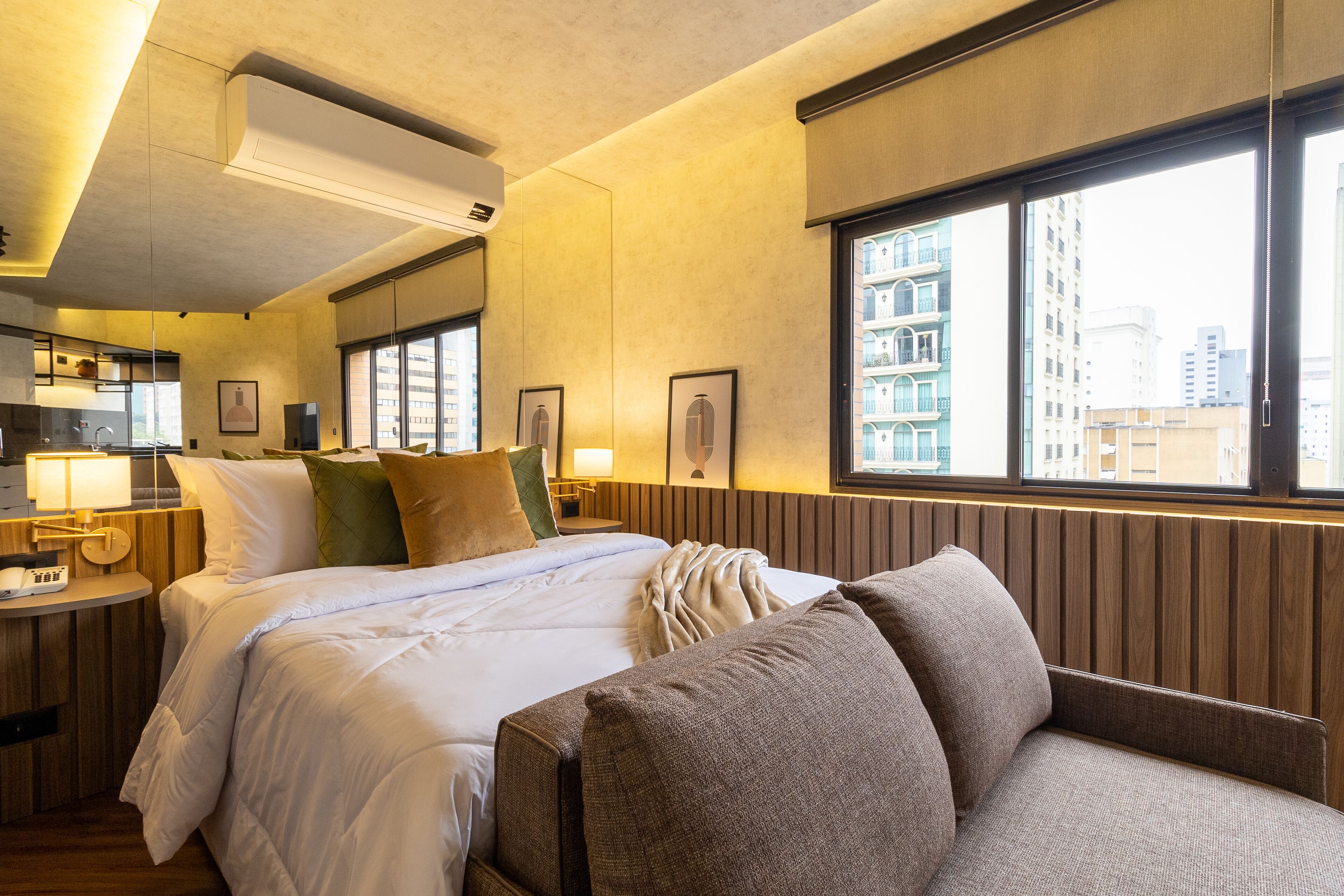 A hotel room with a bed, a sofa, a bedside table with a lamp, a window with a view of buildings, and a wall-mounted air conditioner.