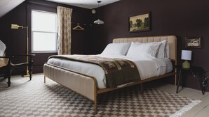 Un dormitorio con suelo a cuadros, una cama con cabecera, una mesita de noche, una lámpara, un aplique y un cuadro en la pared.