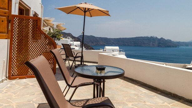 A terrace with a round table and two chairs, a large umbrella, and a view of the sea. - Ostria Caldera Studio 2 (Thira - Santorini)