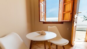 A room with a round table and two white chairs, a window with wooden shutters, and a view of the sea. - Ostria Caldera Studio 2 (Thira - Santorini)