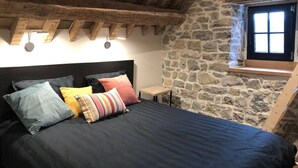 A bedroom with a stone wall, a wooden ceiling, a bed with multiple pillows, and a small table.