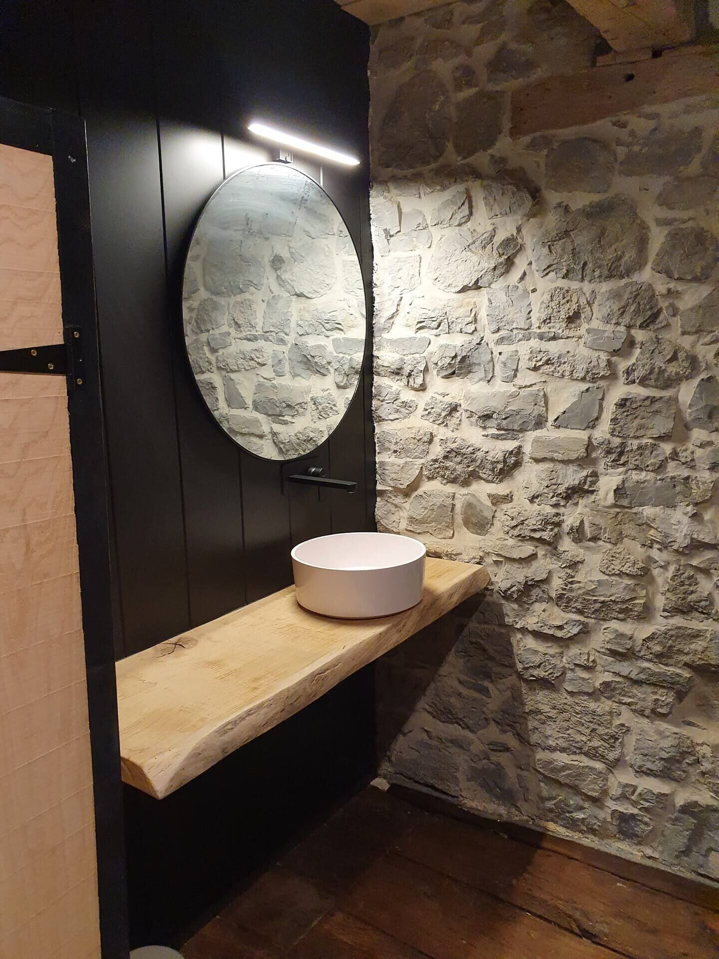 A bathroom with a stone wall, a wooden shelf, a round mirror, and a white sink.