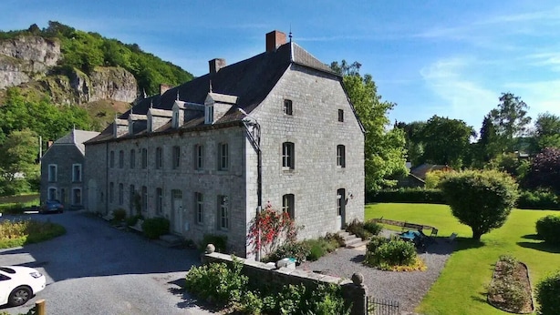 A stone building with a steep roof, a small attached structure, and a well-maintained garden with a lawn and shrubs.