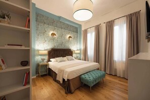 A bedroom with a bed, bedside tables, a lamp, a bench, a TV, and a window with curtains.