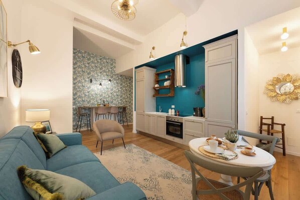 A modern living room with a blue sofa, a dining table with chairs, a kitchen area with teal cabinets, and a patterned rug. - Pepi - Stylish Retreat With Modern Design (Firenze)
