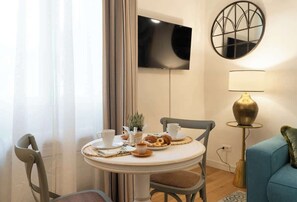 A dining area with a round table, chairs, a TV mounted on the wall, a lamp, and a blue sofa.