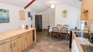 A compact kitchen with wooden cabinets, a dining table with chairs, a refrigerator, and a sink. - The Bookshelf (Swansea)