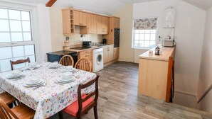 A compact kitchen with wooden cabinets, a washing machine, and a dining area with a floral tablecloth. - The Bookshelf (Swansea)