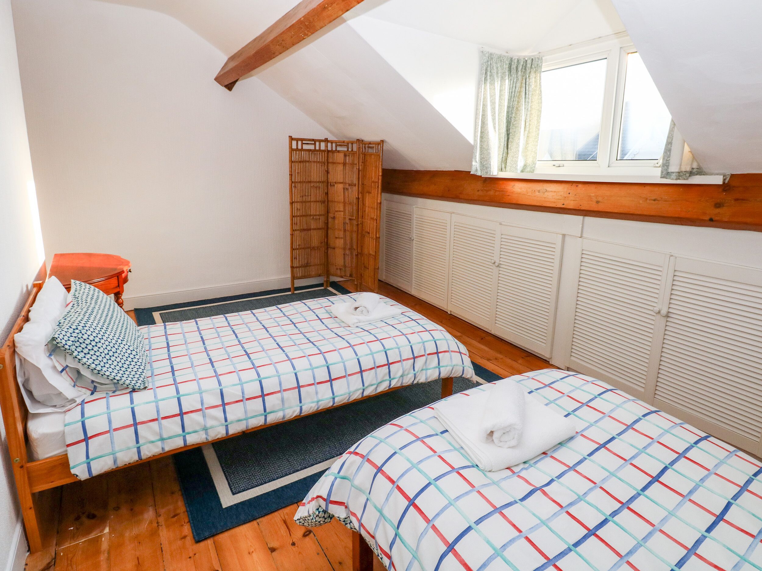 A bedroom with two beds, a window with curtains, a wooden ceiling, and a radiator.