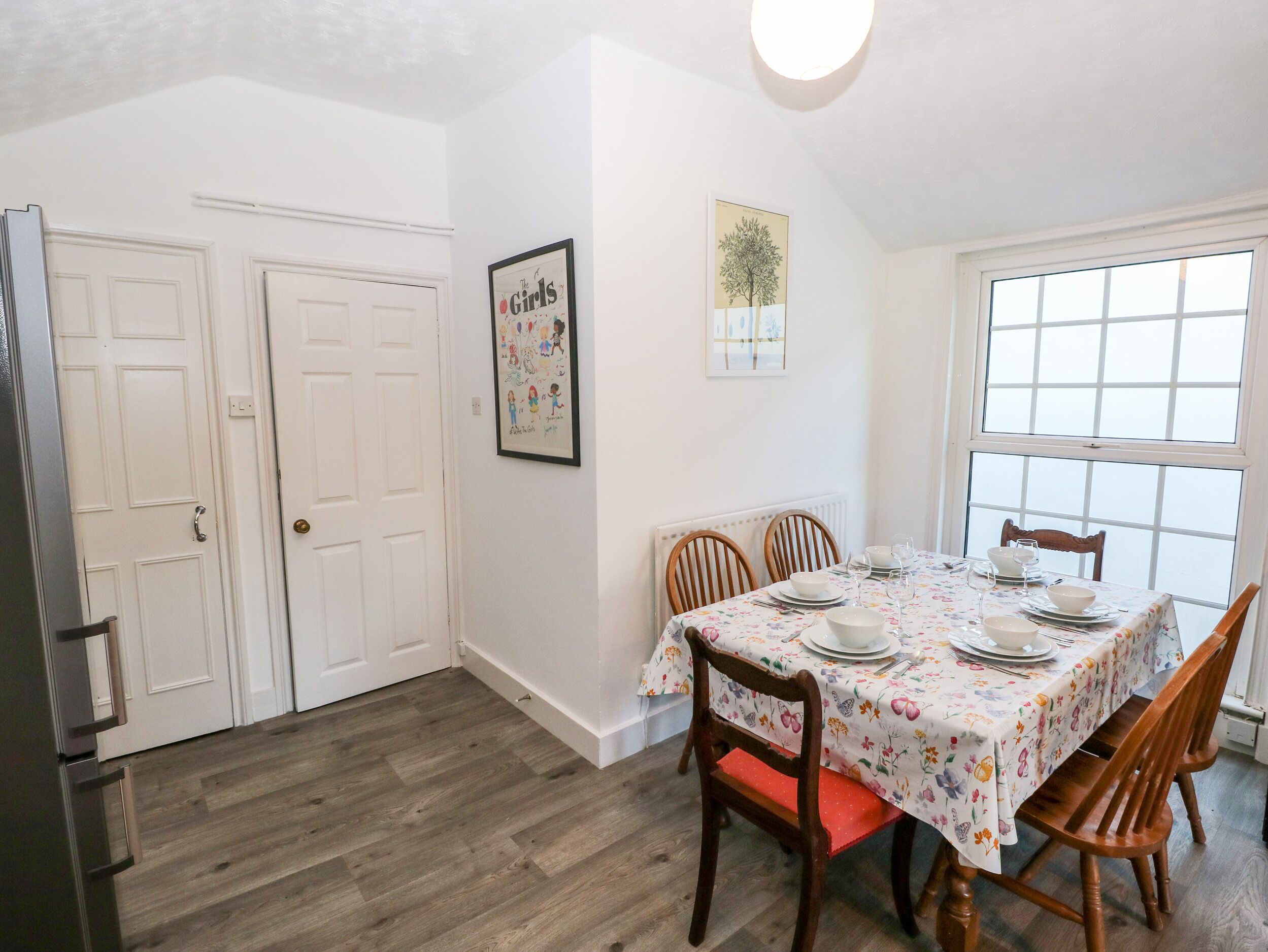 A dining area with a table set for four, wooden chairs, a white door, and a framed picture on the wall.