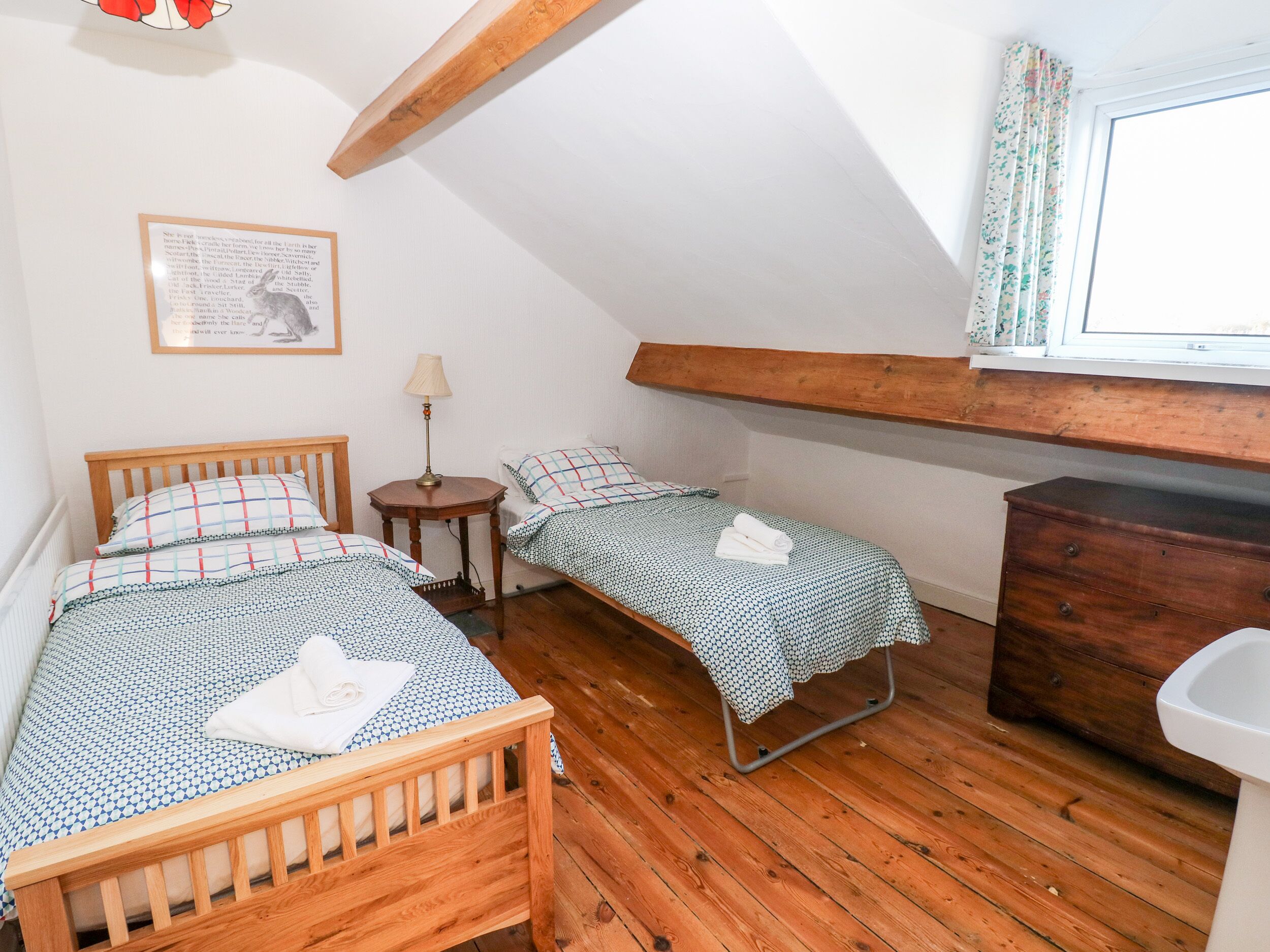 A cozy room with two wooden beds, a small table with a lamp, a dresser, and a window with curtains.