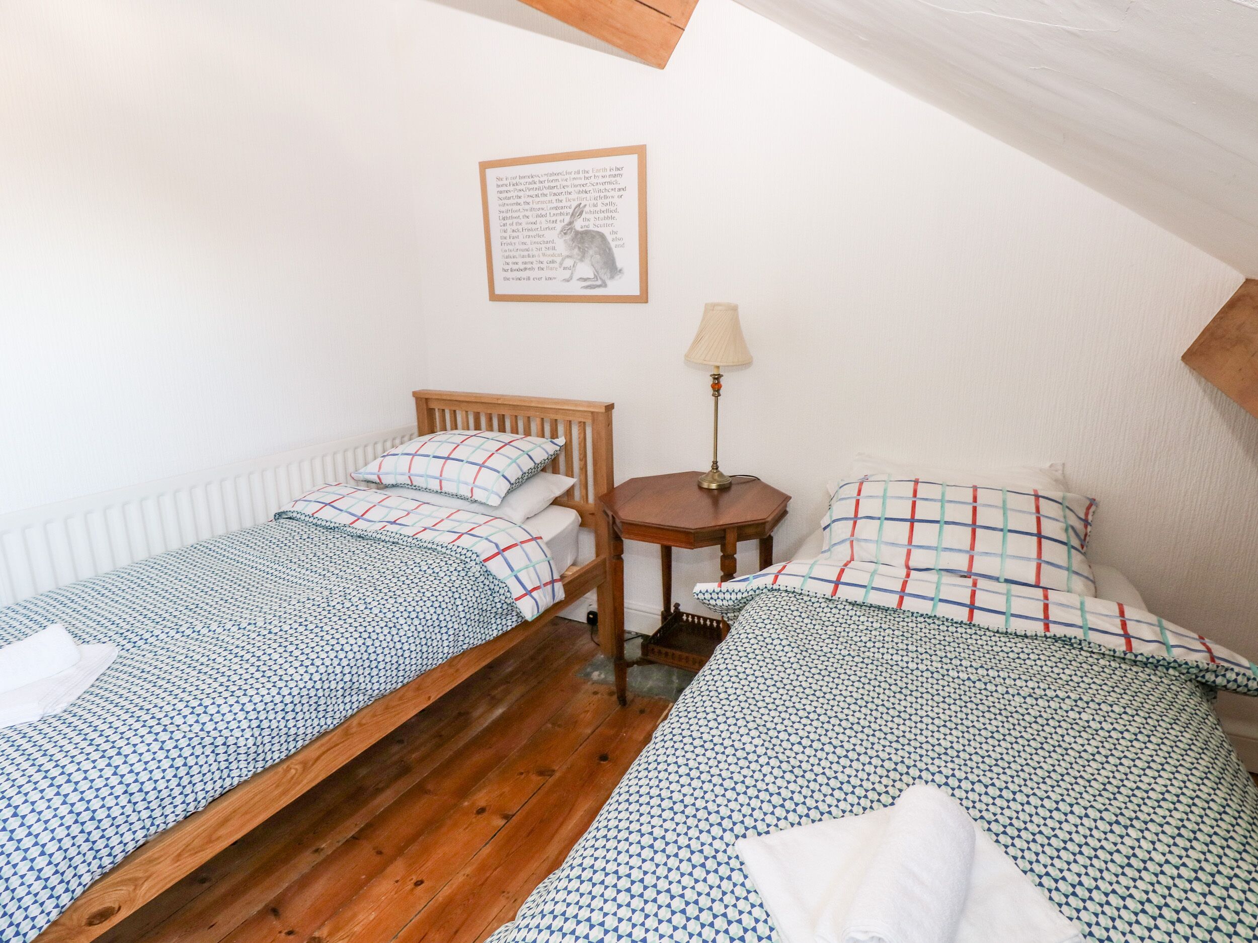 Un dormitorio pequeño y acogedor con dos camas de madera, una mesa redonda con una lámpara y un cuadro enmarcado en la pared.