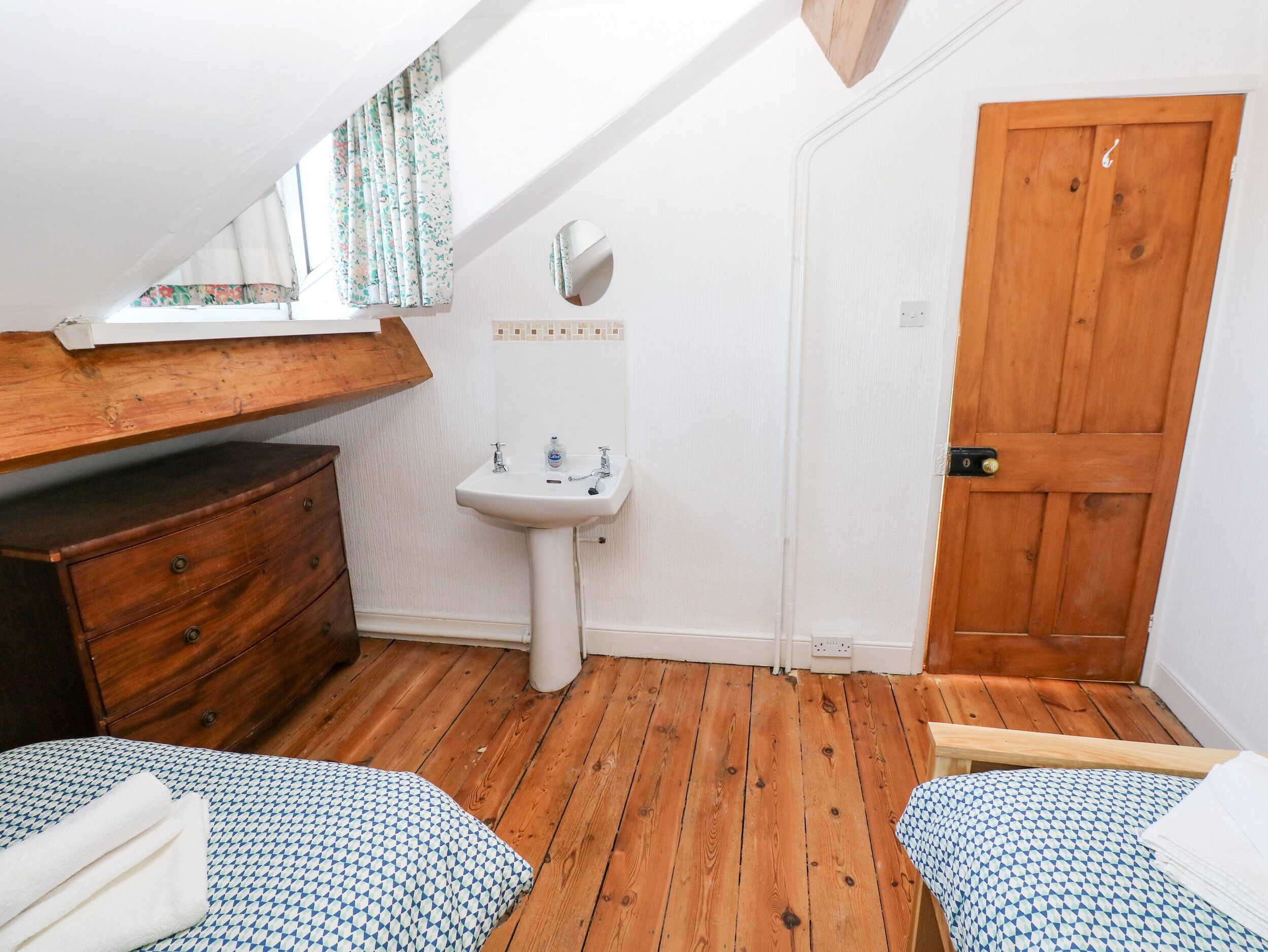 Un dormitorio pequeño y acogedor con suelo de madera, un aparador de madera, un lavamanos blanco y una puerta de madera.