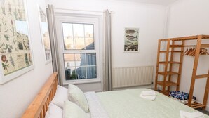 A bedroom with a bed, a wooden headboard, a window with curtains, a radiator, a wooden shelf, and a wall-mounted plant illustration. - The Bookshelf (Swansea)
