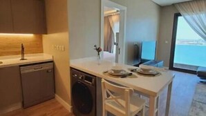 A compact kitchen with wooden flooring, a dishwasher, and a small dining area with a chair. - A Luxurious Sea View 1 Bedroom Apartment (Al HIDD)