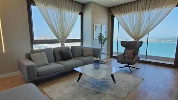A modern living room with a sofa, a coffee table, and a chair. There are large windows with curtains, a plant, and a view of the sea. - A luxurious sea view 2 bedrooms apartment (Al HIDD)
