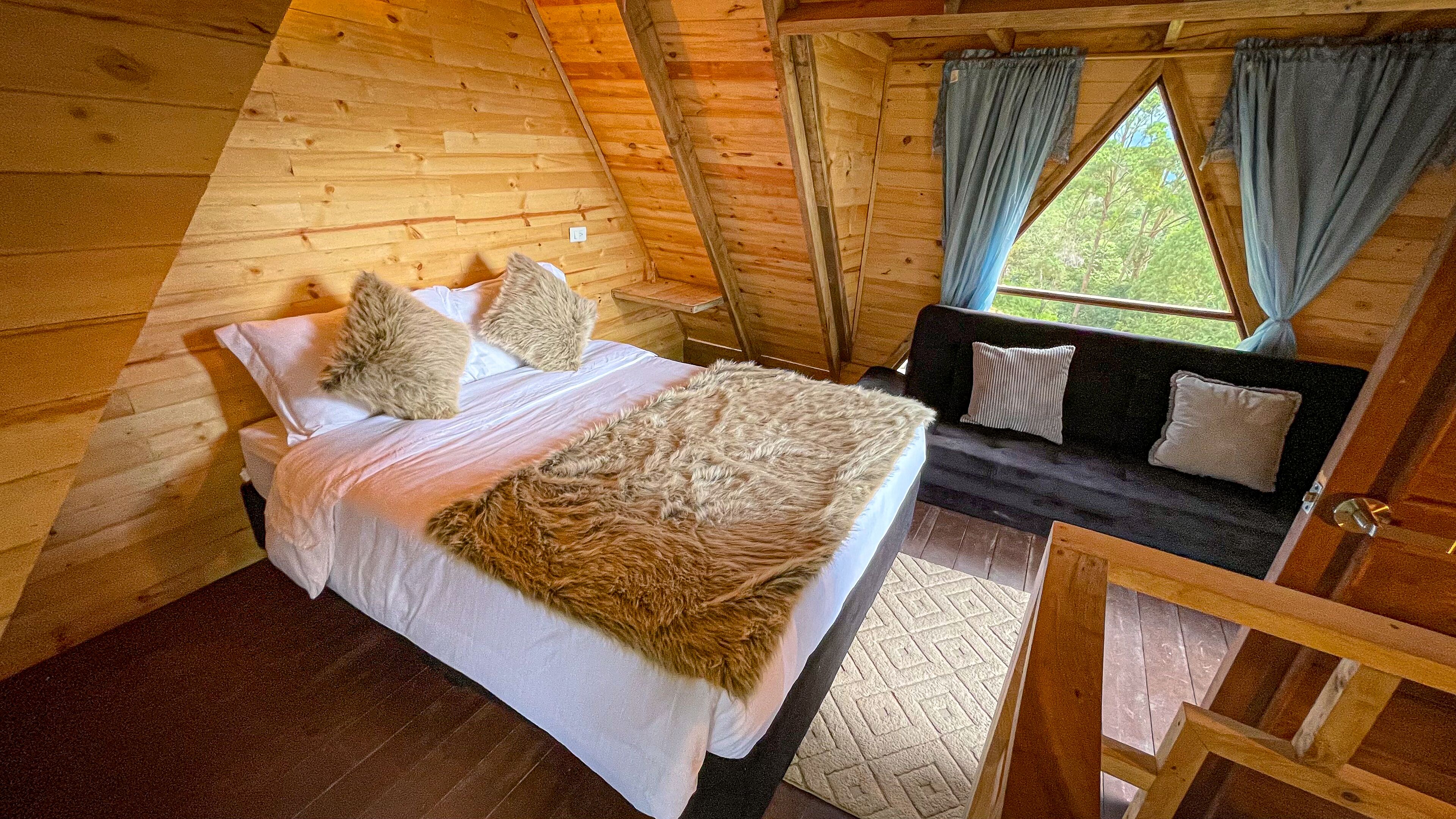 A cozy wooden cabin bedroom with a bed, a sofa, and a window with curtains.