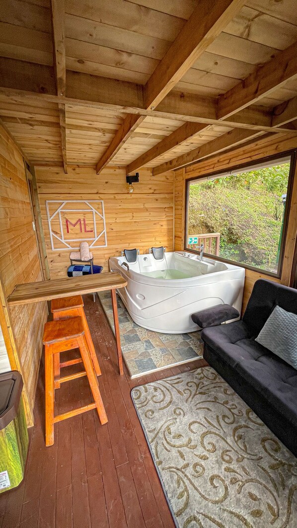 A wooden cabin interior with a large bathtub, a wooden stool, a couch, and a window with a view of greenery. - Glamping Magic Love (San Francisco)