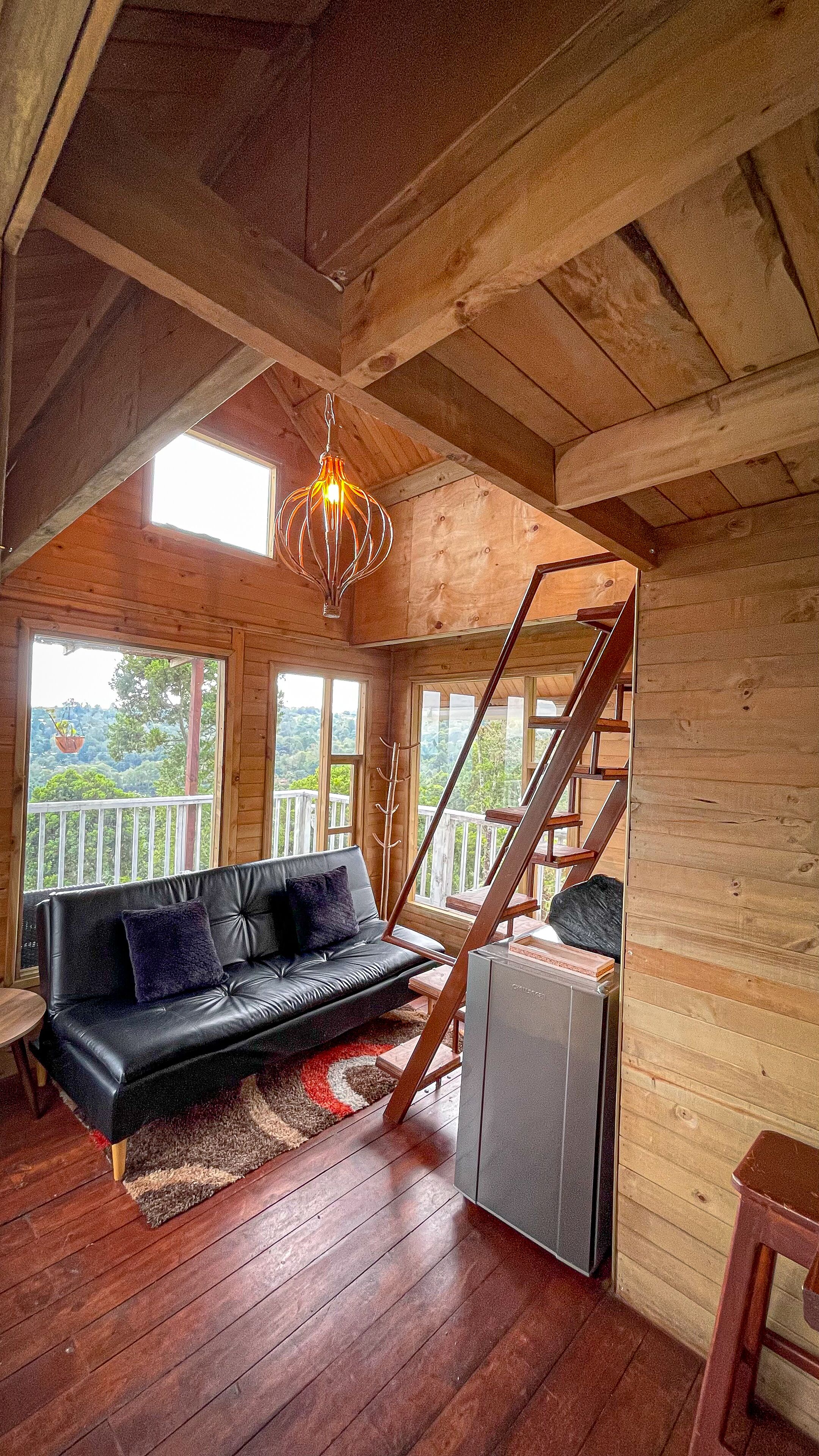 A cozy cabin interior with a wooden staircase, a black leather sofa, and a hanging light fixture.