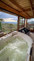 Una cabaña de madera con una bañera grande llena de agua, un sofá con cojines y vistas a un paisaje montañoso a través de amplios ventanales.