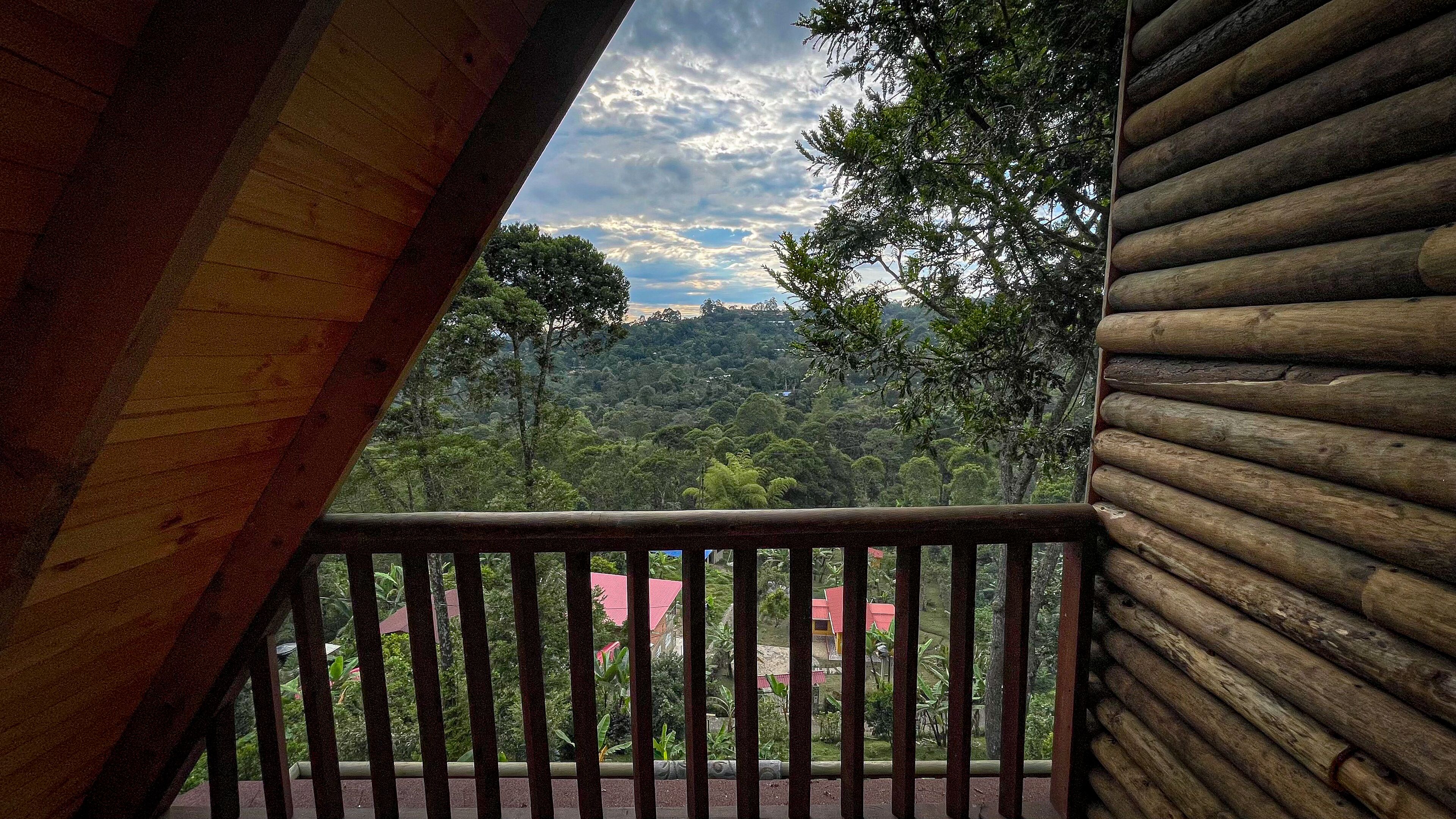 A wooden balcony with a view of a lush green landscape and a red-roofed building in the distance.