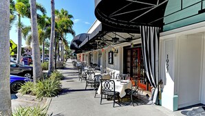 An outdoor dining area with tables and chairs, palm trees, and a building with a curved awning. - Longboat Key Cedars Beach House-pet Friendly-2 Minute To The Beach (Longboat Key)