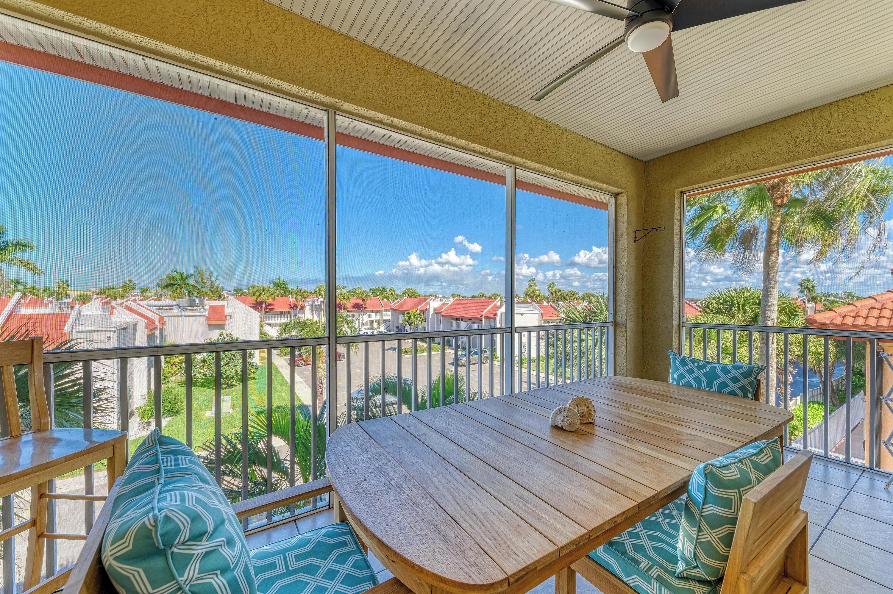 A wooden table with patterned cushions on a balcony with large windows, offering a view of a residential area with red-roofed houses and palm trees.