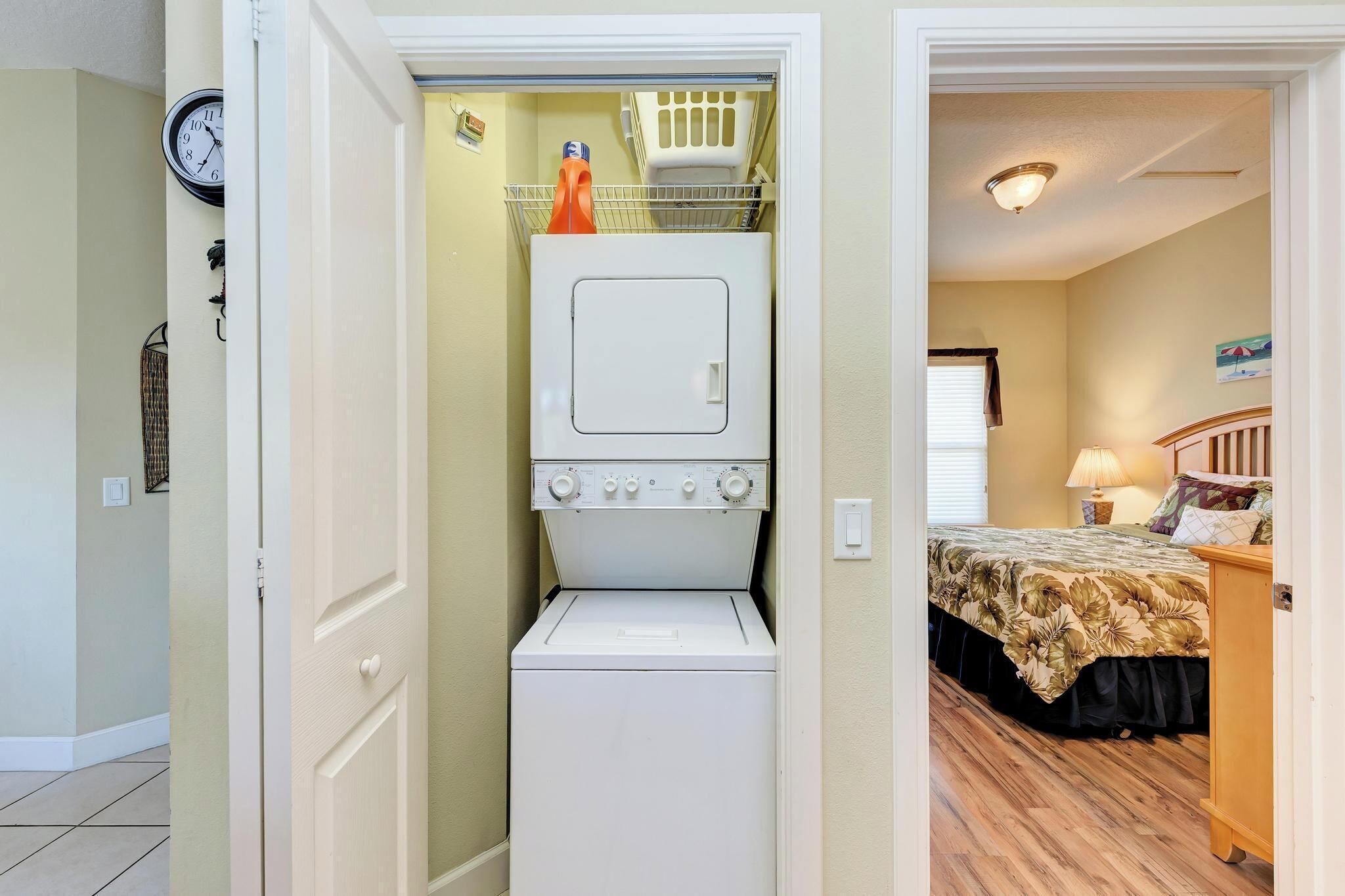 A compact laundry room with a washer and dryer, a clock on the wall, and a bed in the adjacent bedroom.