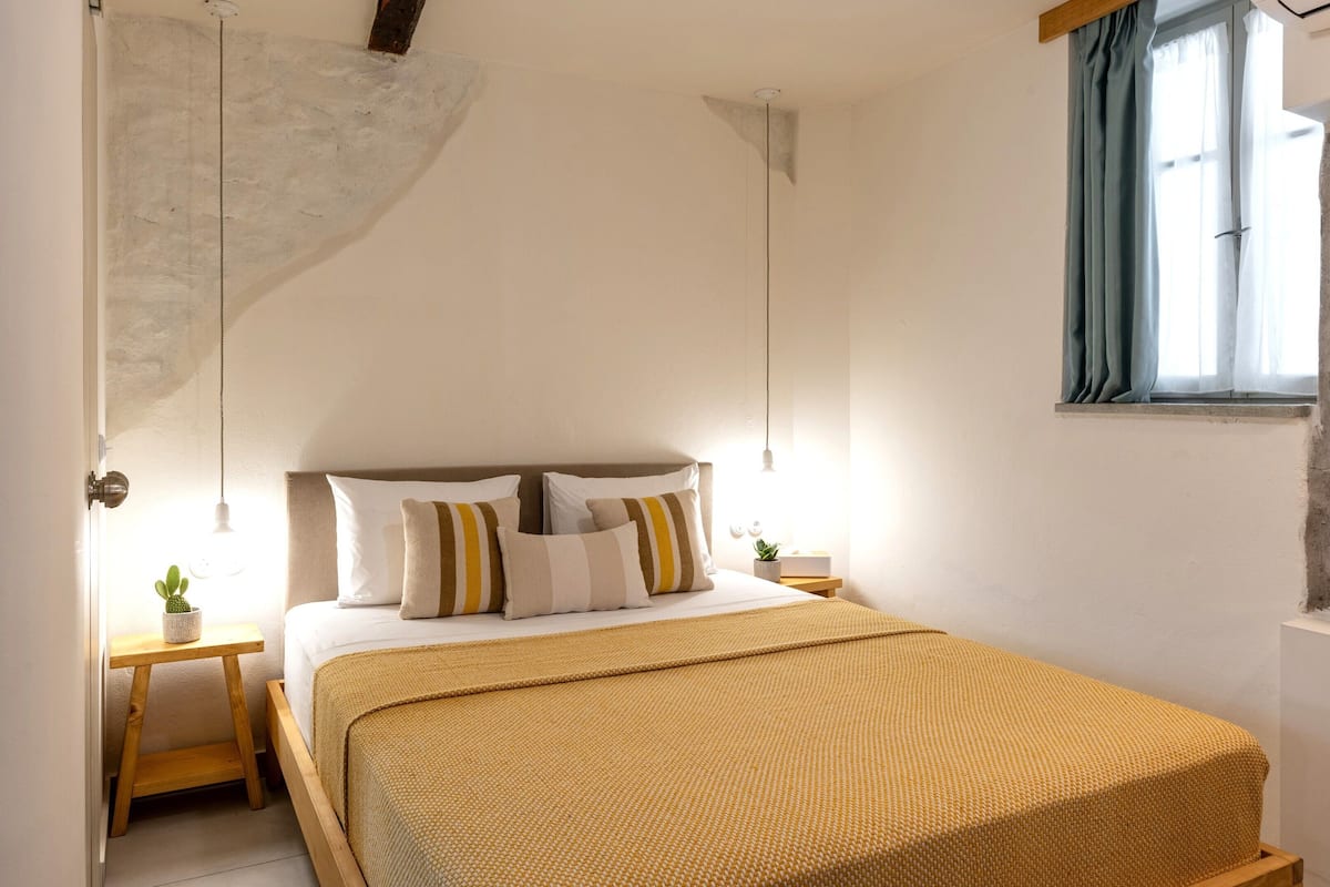 Une chambre à coucher avec un lit, des oreillers, une table de chevet avec une plante, un luminaire suspendu et une fenêtre avec des rideaux.
